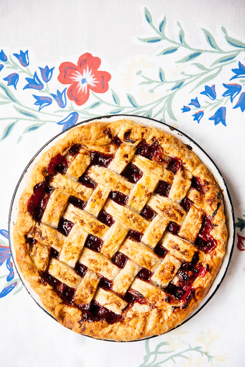 Load image into Gallery viewer, Cherry pie with a lattice crust on a serving plate with a flowered tablecloth.