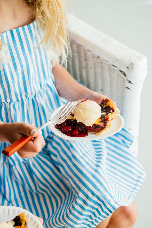 Load image into Gallery viewer, Girl in a blue and white striped dress holding a slice of pie with a fork.