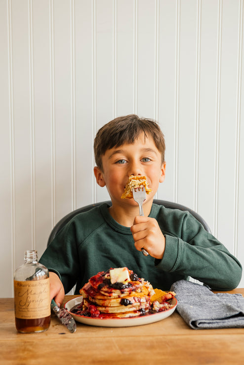 Load image into Gallery viewer, Child eating pancakes, berries and Maple Syrup at a table against a white paneled wall.
