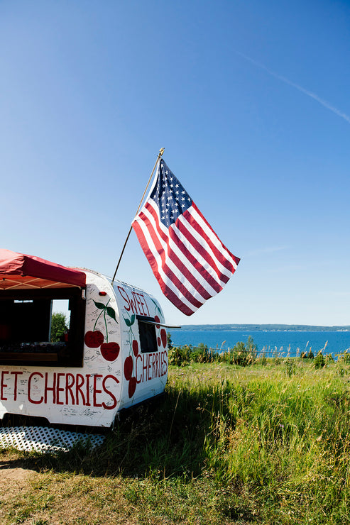 Load image into Gallery viewer, Road side camper cherry stand and American flag in a grassy field with a view of Little Traverse Bay in the background.
