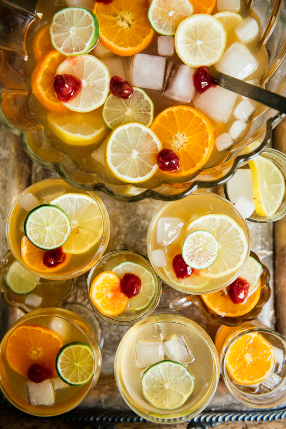 Punch bowl with sliced fruits and ice on a metal tray with served glasses of punch and fruit besides it.