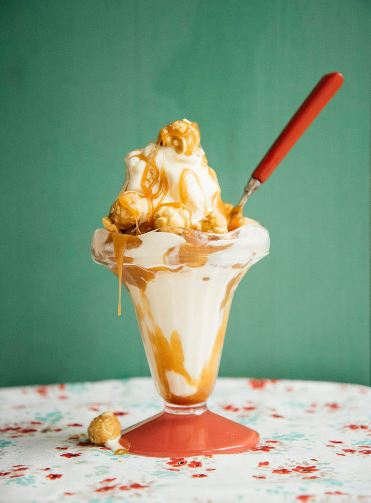 Ice cream sundae with caramel sauce and a red spoon on a floral tablecloth against a green background