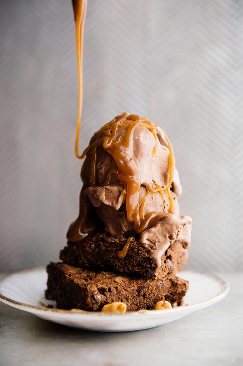 Load image into Gallery viewer, Stack of brownies with ice cream and caramel drizzle on a white plate against a neutral background
