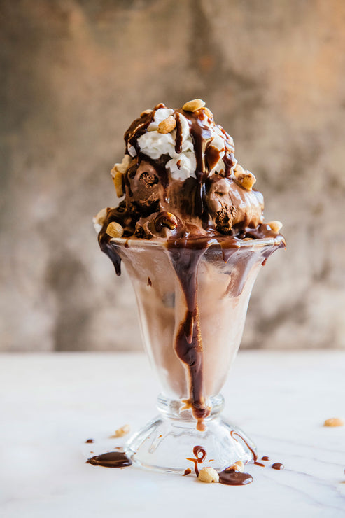 Load image into Gallery viewer, Chocolate ice cream sundae with nuts &amp; whipped cream in a glass cup on a white surface.
