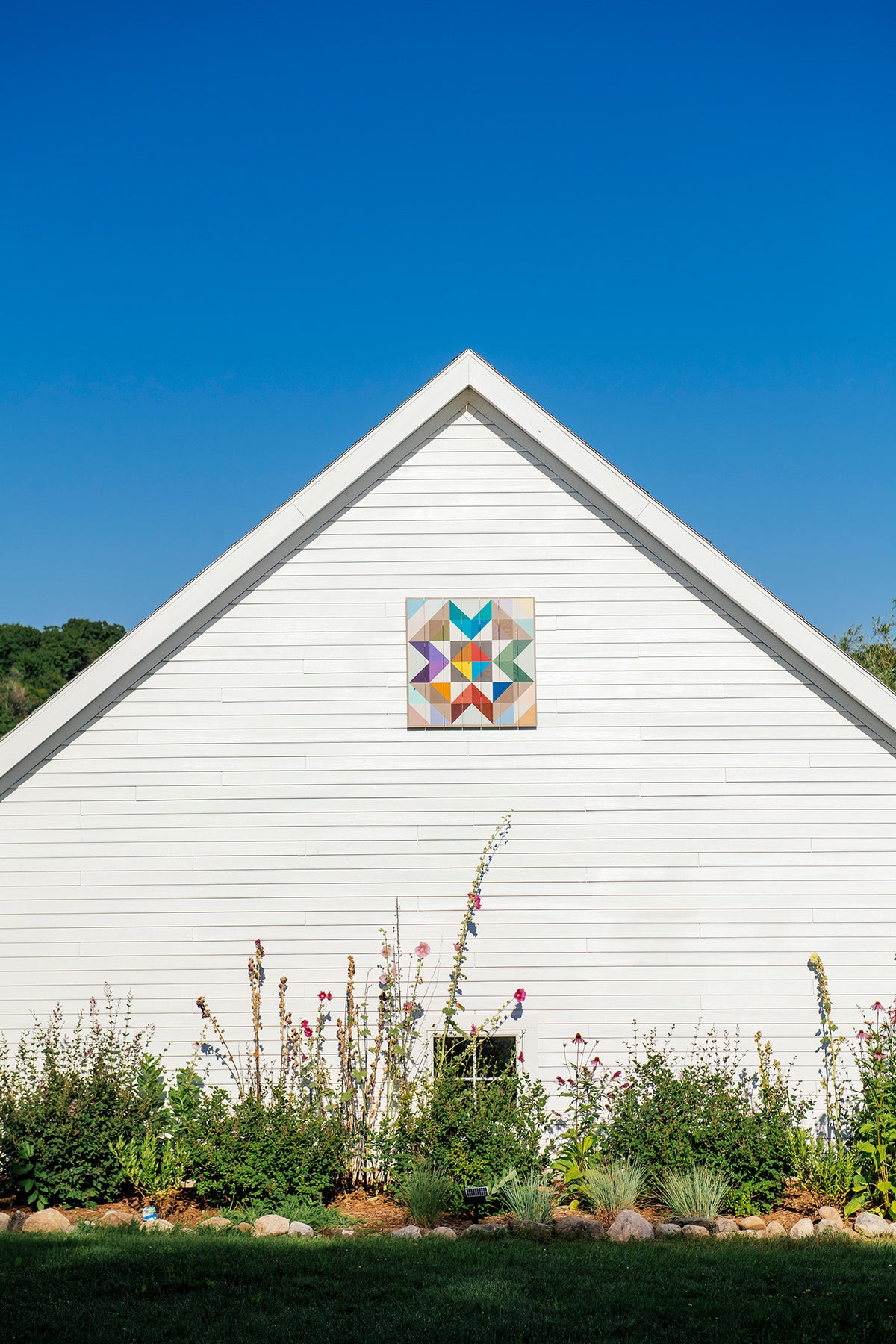 White house with quilt style wooden decor sign and flowers under blue sky.