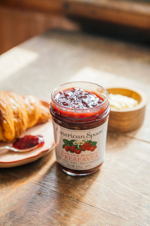 Load image into Gallery viewer, Jar of strawberry preserves on a wooden table with a croissant and a bowl of butter.
