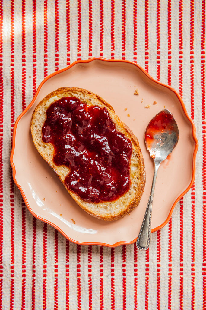 Toast with strawberry preserves on a pink plate with a spoon, all on a red and white striped tablecloth.