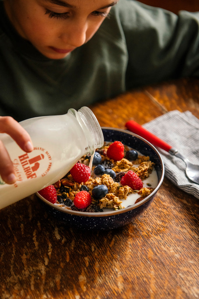 Person pouring milk into a bowl of Maple Granolas with berries on a wooden table