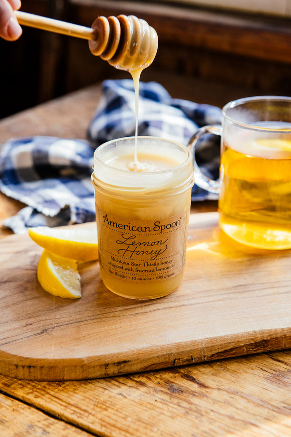 lemon honey being drizzled from a jar with a cup of tea and lemon slices in the background