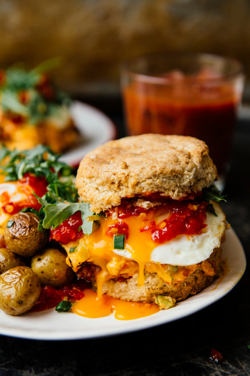 Load image into Gallery viewer, a homemade biscuit breakfast sandwich with potatoes and arugula on the side topped with chili jam.
