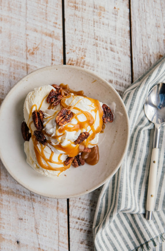 Ice cream with caramel and pecans on a white bowl, placed on a wooden surface with a striped cloth and spoon.