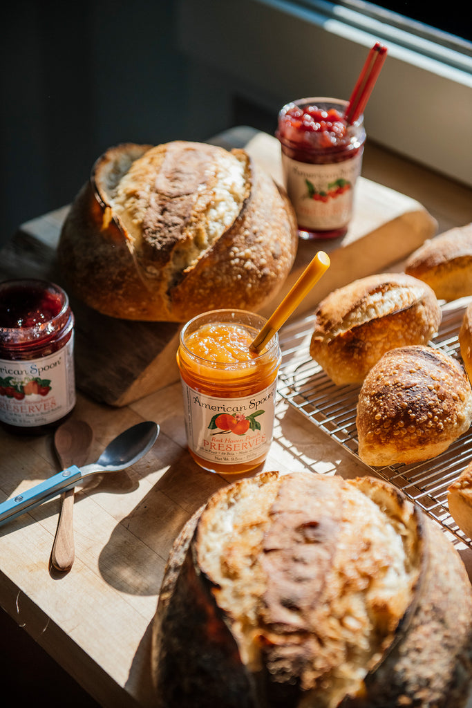 Loaves of bread with jars of Peach, Strawberry & Sour Cherry Preserves on a wooden surface