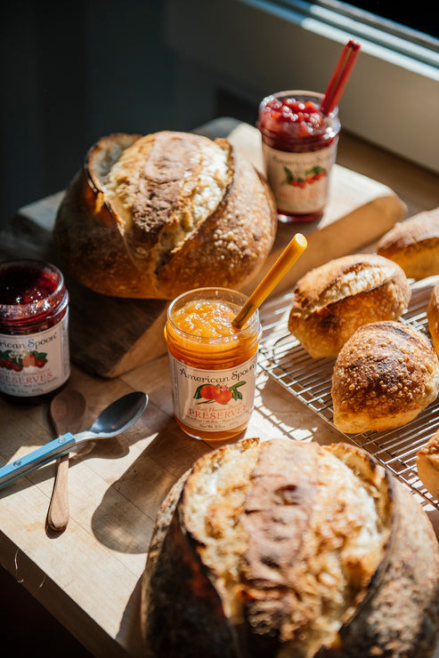 Load image into Gallery viewer, Loaves of bread with jars of Peach, Strawberry &amp; Sour Cherry Preserves on a wooden surface
