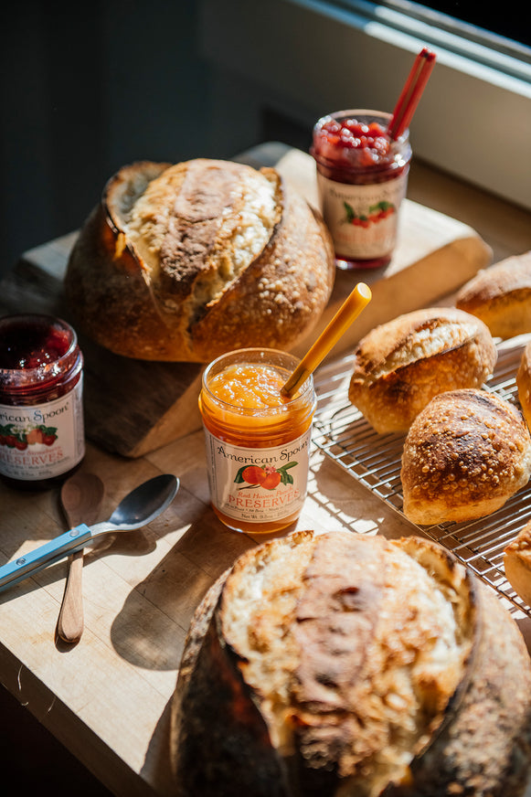 Loaves of bread with jars of Peach, Strawberry & Sour Cherry Preserves on a wooden surface
