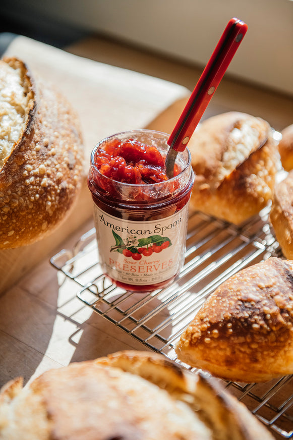 Jar of American Spoon Sour Cherry Preserves with a spoon on a cooling rack with bread