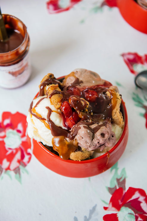 Load image into Gallery viewer, Ice cream sundae with chocolate sauce, cherries, and cookies in a red bowl on a floral tablecloth.
