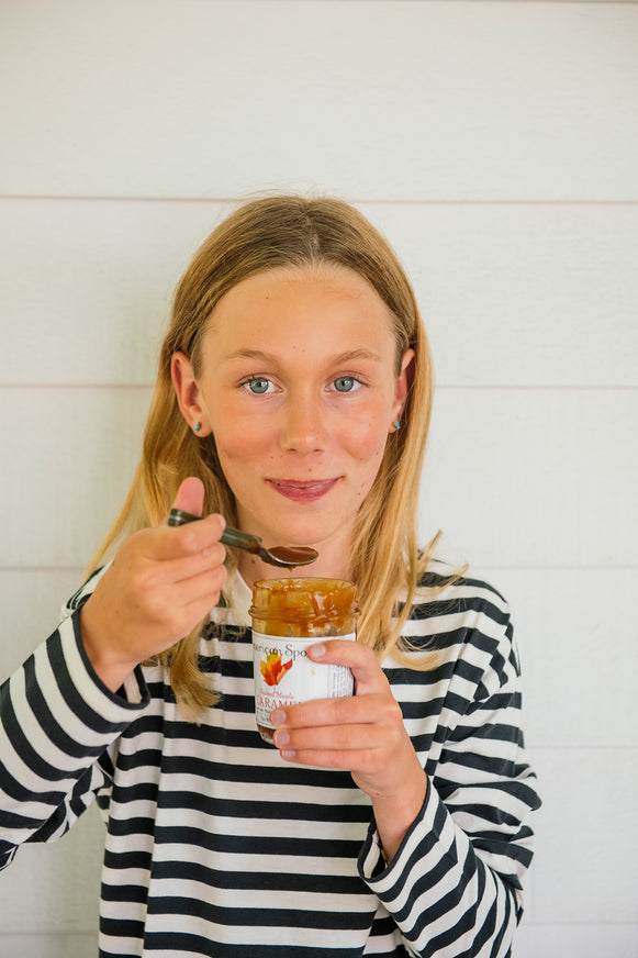 Young girl holding a jar of Salted Maple Caramel and a spoon against a white background