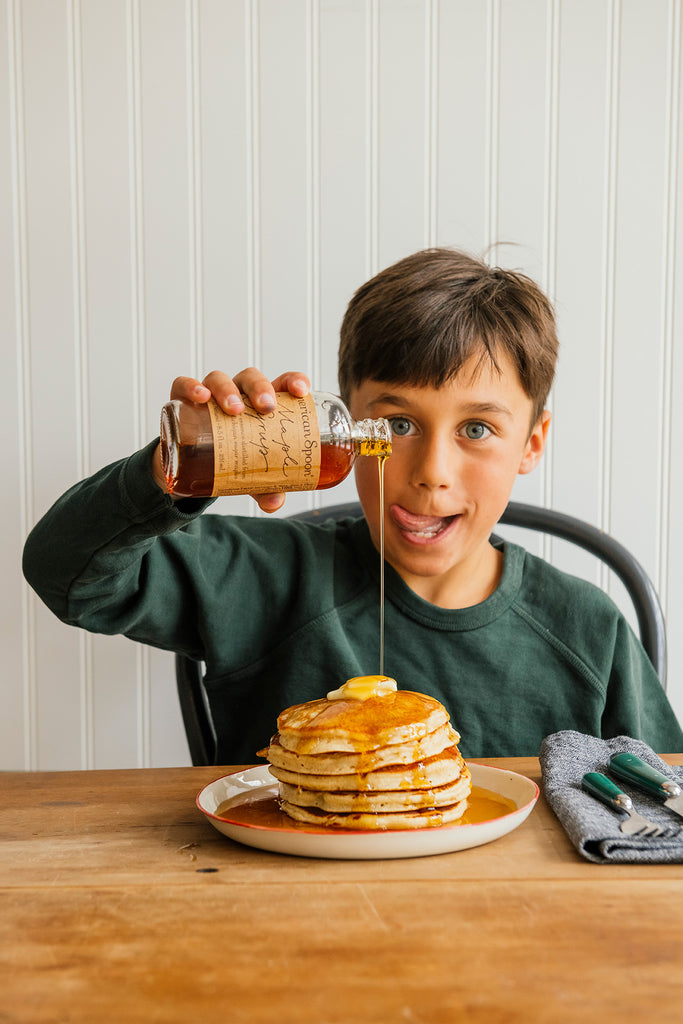 Child pouring syrup onto a stack of pancakes with berries on a wooden table.