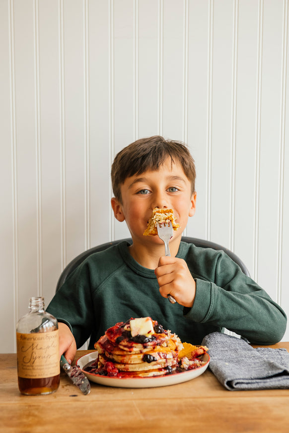 Child eating pancakes, berries and Maple Syrup  at a table against a white paneled wall.