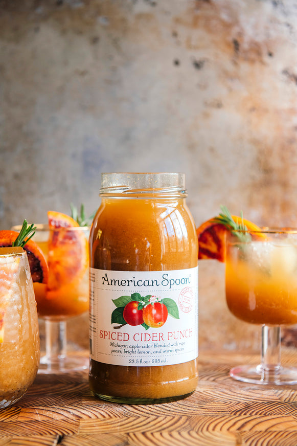 Jar of American Spoon Spiced Cider Punch on a wooden surface with glasses of cider in the background.
