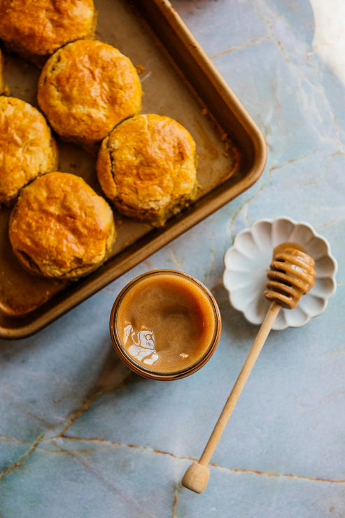 Load image into Gallery viewer, Biscuits on a sheet pan with a jar of Cinnamon Honey and a honey dipper on a marble surface.
