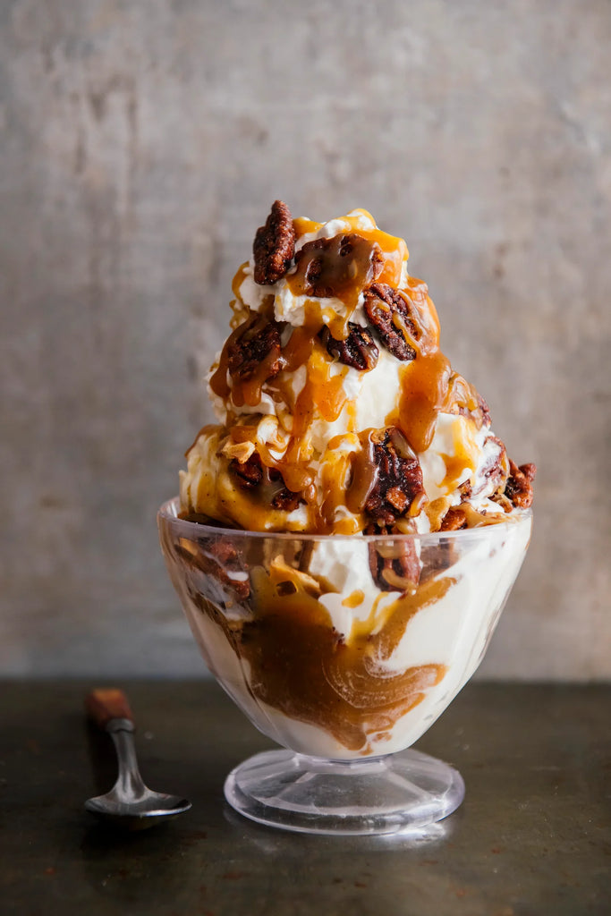 Dessert with ice cream, caramel, and pecans in a glass bowl on a textured surface.