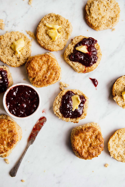 Load image into Gallery viewer, Overhead of several golden-brown baked biscuits on a white marble surface, with some sliced open and topped with butter and strawberry preserves A small white bowl of jam and a knife are also visible.
