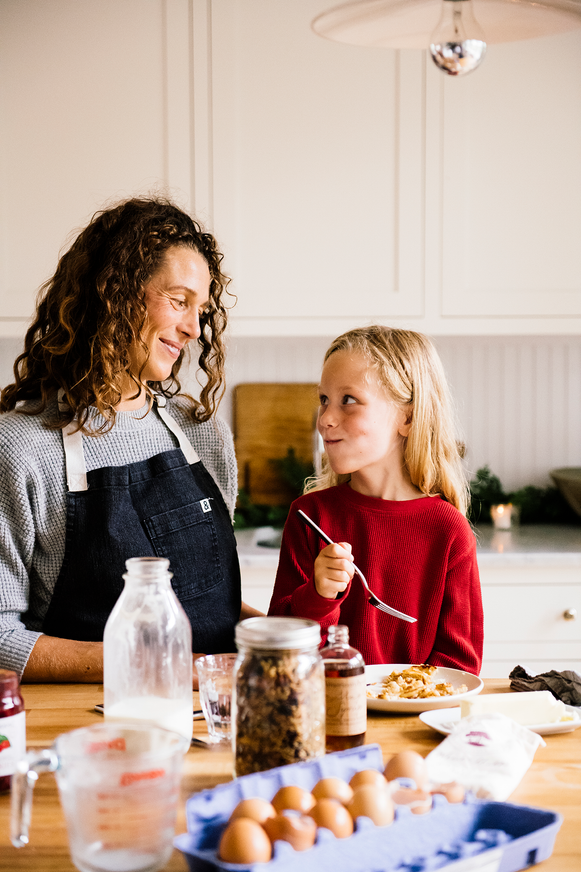 a mother and daughter eating pancakes at a kitchen island with various breakfast ingredients