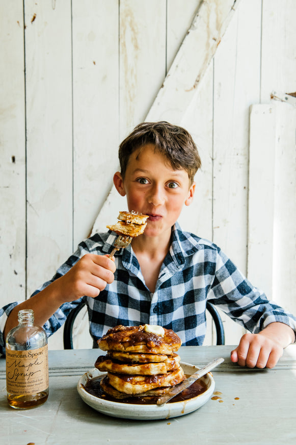 A young boy with a fork full of pancakes and a stack of pancakes covered in Maple Syrup